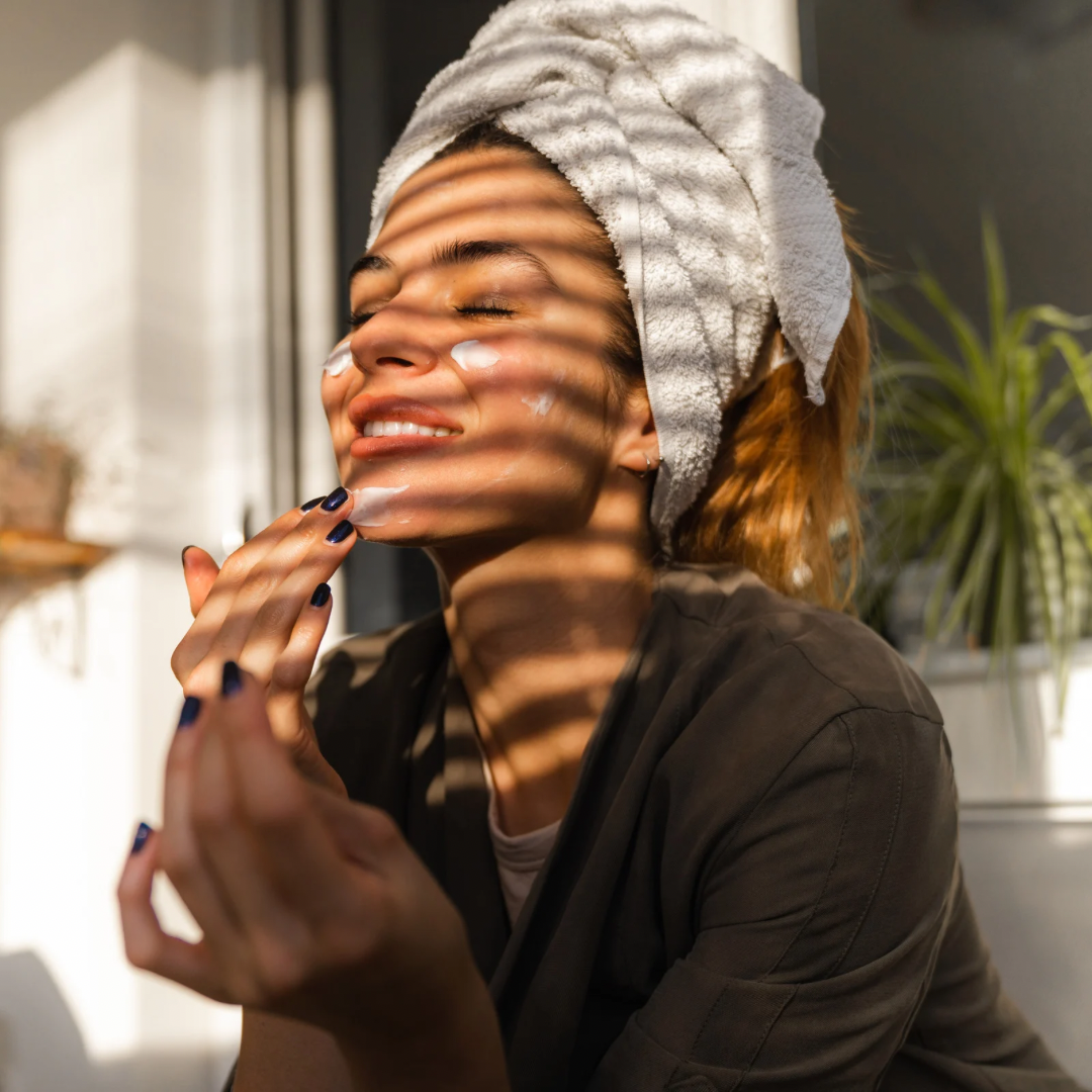 Smiling woman relaxing with a towel wrapped around her head, enjoying a self-care moment in soft natural lighting