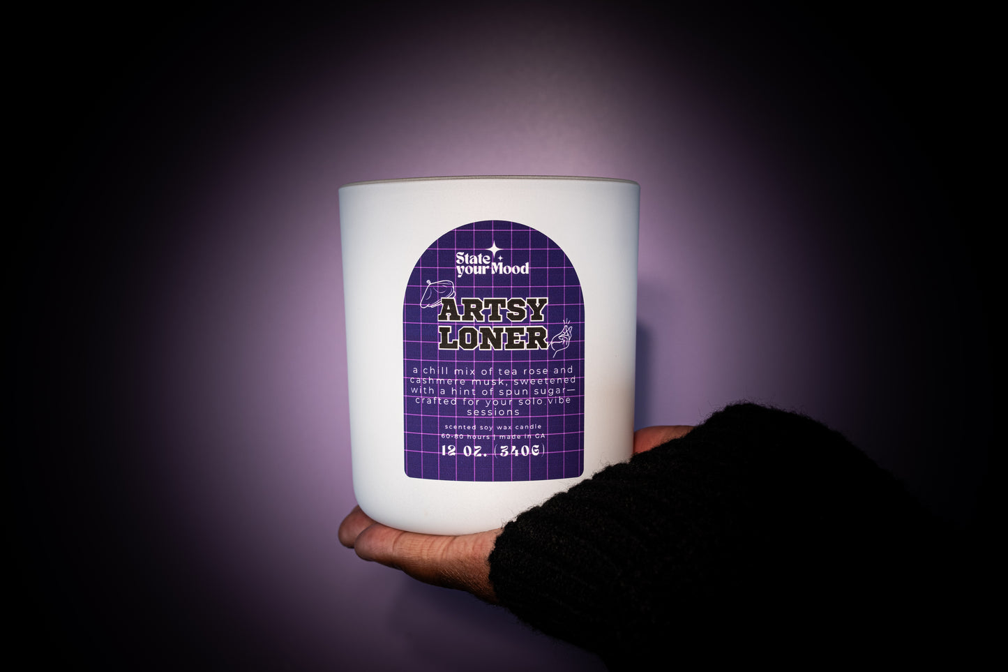 A hand holding the 'Artsy Loner' candle from State Your Mood. The candle is a white matte jar with a purple grid-patterned label and bold text. The hand is wearing a dark, cozy sweater, and the background is a soft gradient of purple lighting that creates a spotlight effect on the candle, emphasizing its minimalist and artistic vibe. The candle size is noted as 12 oz (340g).