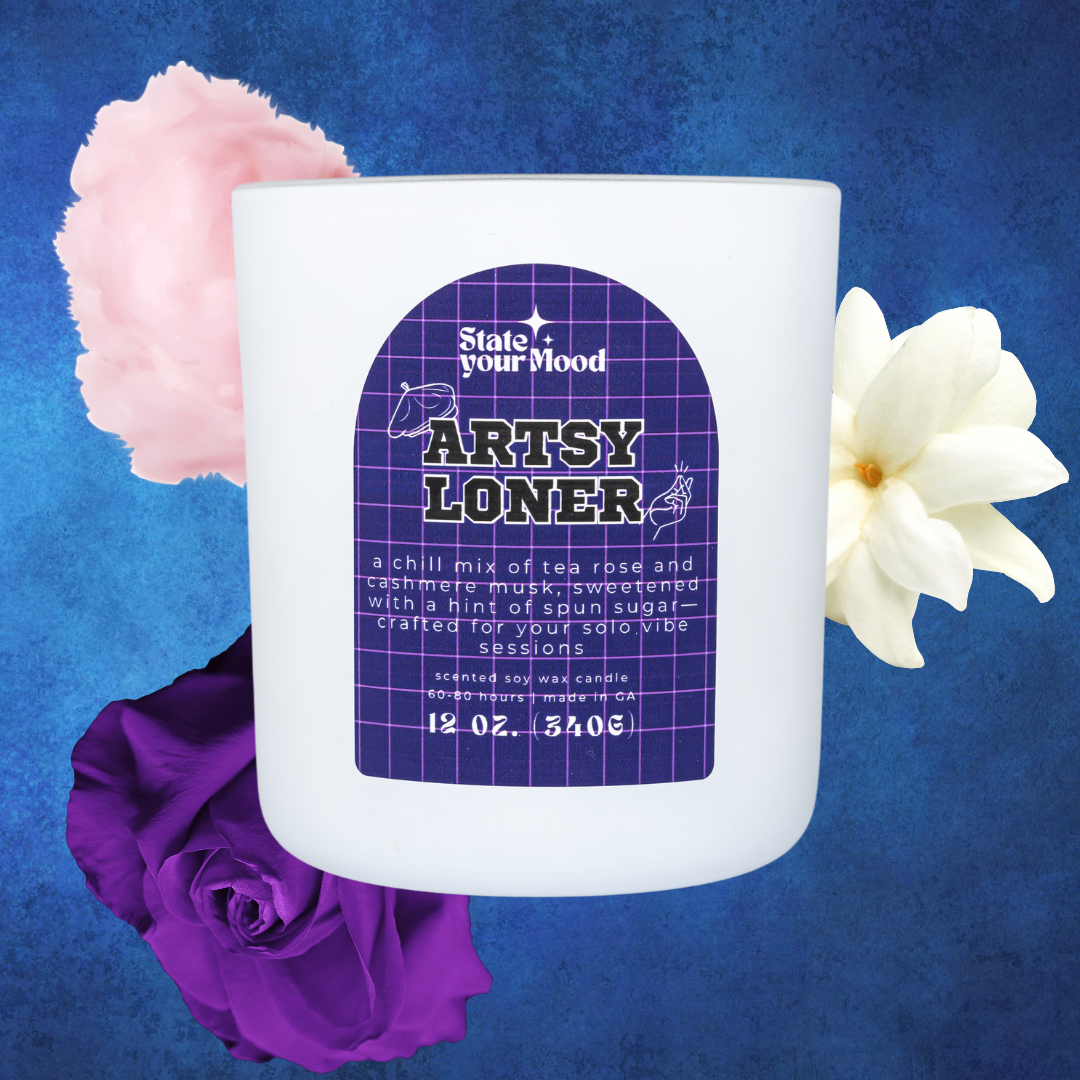 A white matte candle jar labeled 'Artsy Loner' from the State Your Mood brand, featuring a purple grid-patterned label with bold text. The description highlights the scent as a 'chill mix of tea rose and cashmere musk, sweetened with a hint of spun sugar.' Surrounding the candle are soft floral accents, including a pale pink peony, a white gardenia, and a deep purple rose, set against a textured blue background, enhancing the serene and introspective vibe.