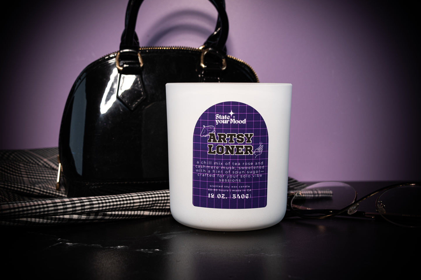 The 'Artsy Loner' candle by State Your Mood placed on a sleek black surface. The white matte jar with a purple grid-patterned label is in the foreground. Behind the candle is a stylish black handbag, a pair of glasses, and a plaid skirt draped casually, creating a moody and artistic vibe. The background features soft purple lighting that adds to the minimalist, introspective theme of the setup.