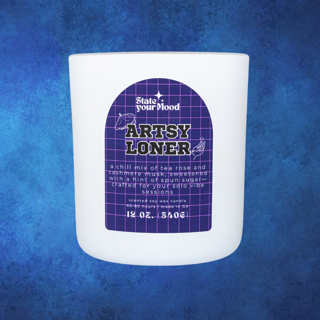 A white matte candle jar labeled 'Artsy Loner' from the State Your Mood brand. The label has a purple grid pattern, with the candle's name in bold, retro-inspired font. The description reads: 'A chill mix of tea rose and cashmere musk, sweetened with a hint of spun sugar—crafted for solo vibe sessions.' The candle size is listed as 12 oz. (340g). The background is a textured blue, complementing the minimalistic and artistic vibe of the candle.