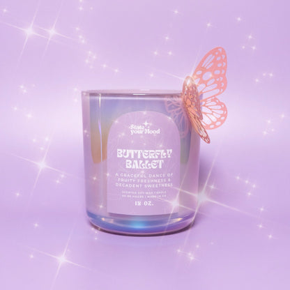 A delicate, iridescent candle jar with a pastel purple hue, labeled 'Butterfly Ballet' from the State Your Mood brand. A small, intricate orange butterfly decoration is attached to the side of the jar, giving the illusion of it landing on the candle. The label reads: 'A graceful dance of fruity freshness & decadent sweetness.' The background is a soft lavender with sparkly, twinkling effects, creating a magical and whimsical atmosphere. The candle size is noted as 12 oz.