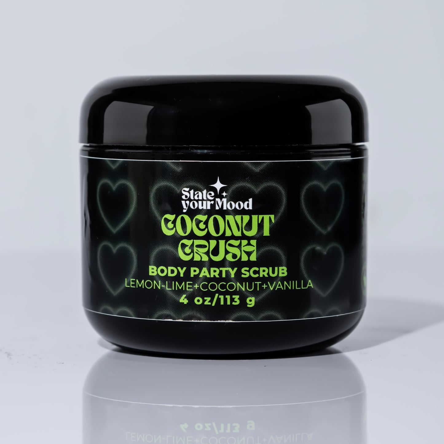 A black jar of 'Coconut Crush Body Party Scrub' by State Your Mood. The label features neon green text with the scent description 'Lemon + Lime + Coconut + Vanilla.' The background of the jar is decorated with soft, heart-shaped outlines, giving the design a playful and vibrant vibe. The jar contains 4 oz (113g) of body scrub, and its reflection is visible on the glossy white surface beneath, creating a sleek and clean look.