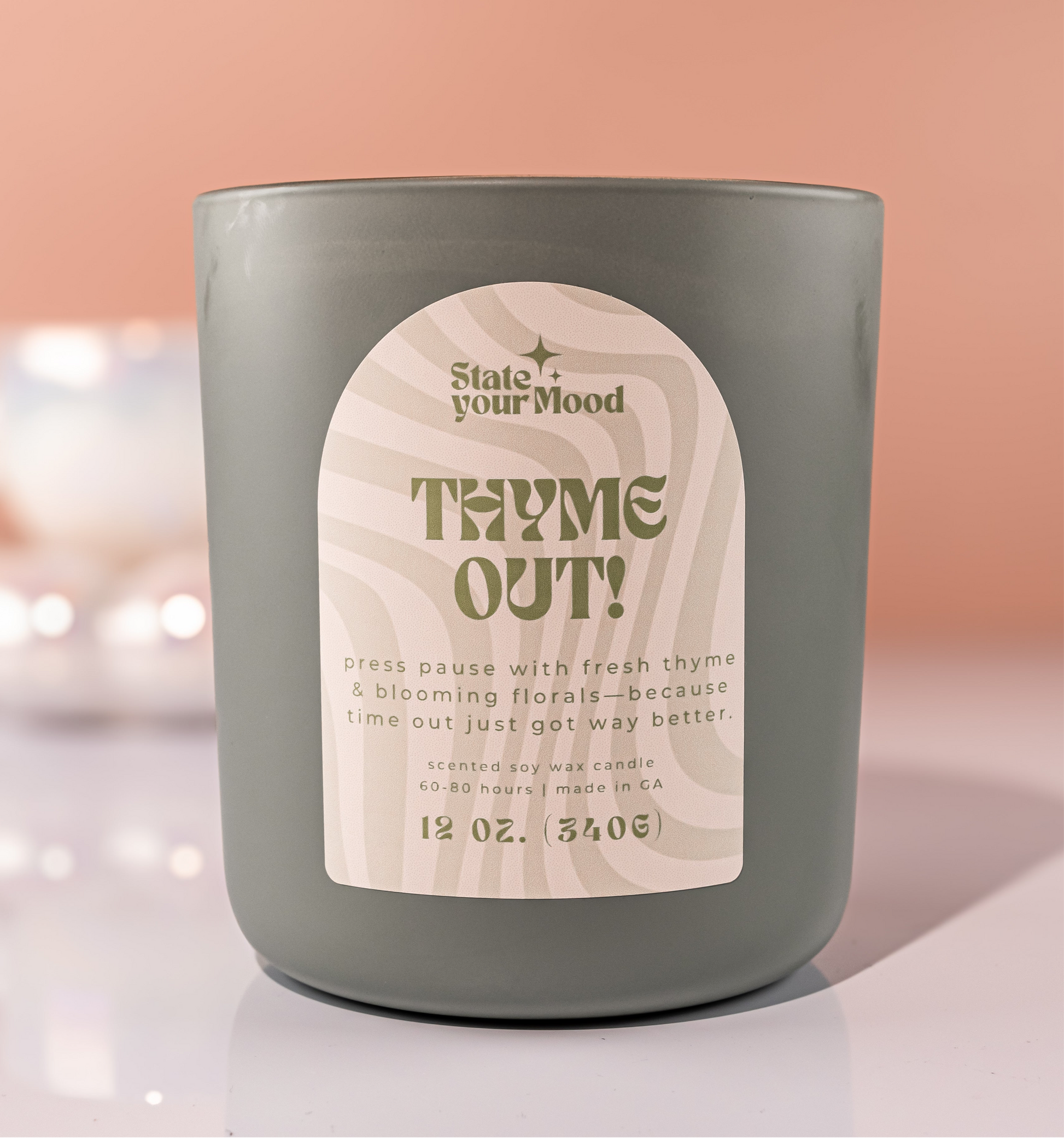 A matte olive-green candle jar with a wavy beige label displaying the text 'Thyme Out!' in bold, retro-style font. Below the title, the description reads: 'Press pause with fresh thyme & blooming florals—because time out just got way better.' The 'State Your Mood' logo is placed above the title, and the candle size '12 oz. 340g' is listed at the bottom. The background features a soft, warm setting with blurred lights, creating a calm and cozy ambiance.